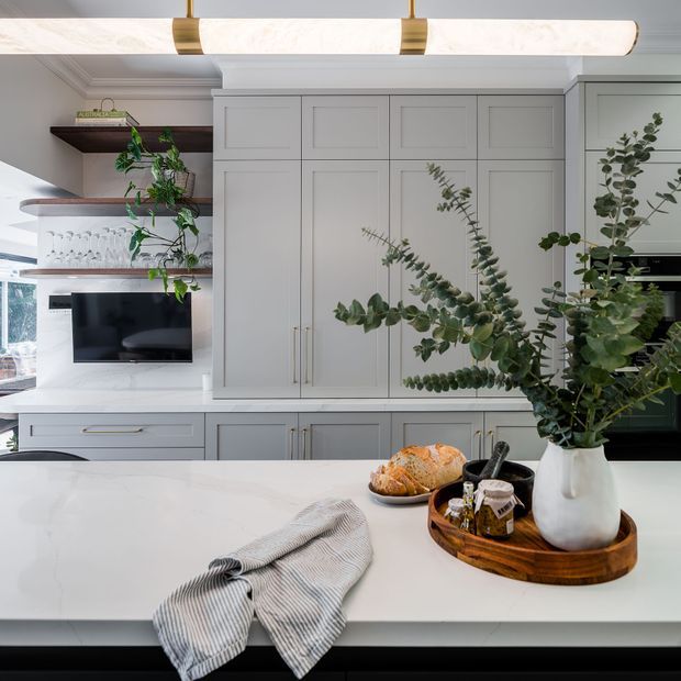 Bright, refined kitchen interior with extensive pale grey built-in shaker cabinetry, a large white island benchtop, open timber shelving, integrated appliances, and a sculptural linear pendant light. A timber tray with a white vase and eucalyptus branches provides an organic focal point, while a small seating and living area are visible beyond the kitchen.