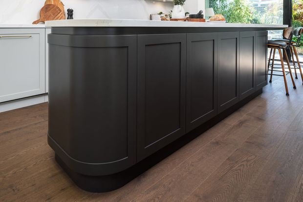 A bright, refined kitchen centred on a large dark charcoal island with framed panel cabinetry and rounded ends. The island has a light stone benchtop, while the surrounding cabinetry is pale grey or white. Warm timber flooring, dining stools, and natural light create a welcoming contemporary-traditional feel.