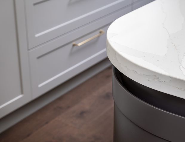 Close-up detail of a contemporary kitchen, showing a rounded white veined stone benchtop over a dark curved island base, with grey shaker-style cabinetry, brass handles, and dark timber flooring.