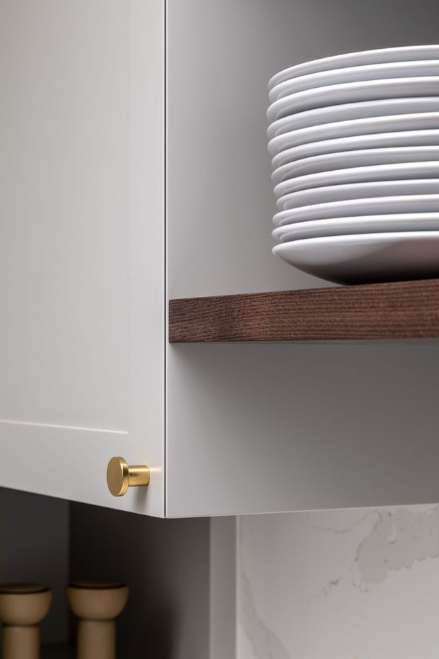 Close-up detail of a contemporary built-in kitchen or dining-area cabinet in a soft white finish, with a brushed brass round knob, dark timber open shelf, stacked white ceramic plates, and a pale veined stone surface below. The composition highlights refined joinery, warm wood, brass hardware, and neutral finishes.