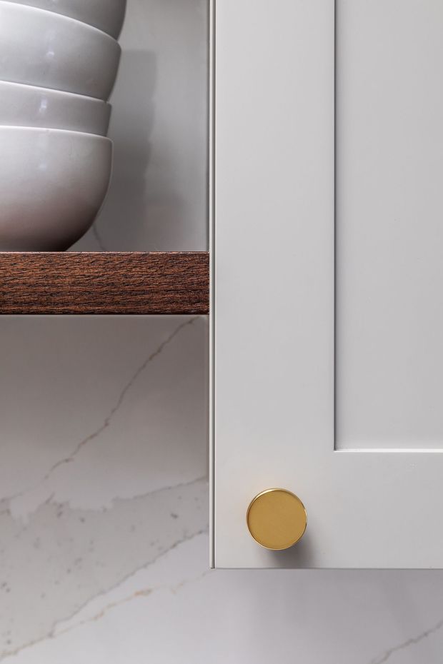 Close-up detail of refined built-in cabinetry featuring a white painted shaker-style door with a round brass-gold knob, a dark timber shelf, white ceramic vessels, and a white marble-look stone surface with subtle veining.