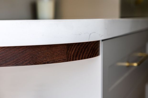 Close-up detail of contemporary built-in cabinetry, showing a white smooth benchtop or cabinet edge, white drawer fronts, and a distinctive dark reddish-brown timber-grain inset handle or drawer pull. A blurred brass-toned handle is visible on adjacent cabinetry in the background.