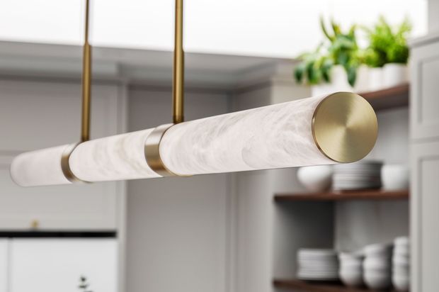 Close-up of a contemporary linear pendant light with a white marble-look cylindrical diffuser and brushed brass end caps and suspension rods. It hangs over a refined kitchen or dining area, with white cabinetry and dark wood open shelving displaying crockery in the background.