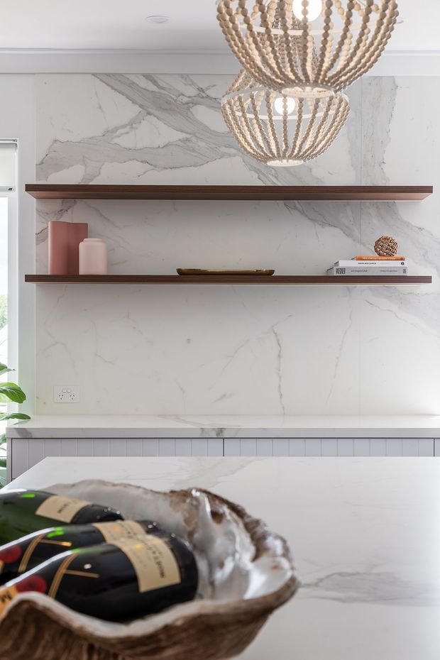 A refined kitchen or entertaining space featuring a dramatic white marble-look slab backsplash and matching island surface, dark timber floating shelves, and warm woven pendant lights. Decorative vessels, books, and a sculptural bowl add styling, while a champagne-and-ice arrangement sits in the foreground.
