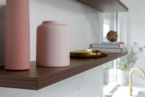 Close-up of a refined kitchen or living-area built-in display shelf in dark timber, set against a pale veined stone or marble-look wall. The shelf holds sculptural ceramic vessels, books, and a brass decorative bowl, with a glimpse of a bright kitchen workspace and gold-toned curved tap in the background. The composition is warm, minimal, and contemporary.