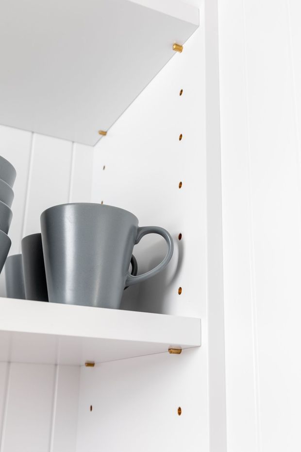 Close-up of white built-in open shelving with adjustable shelf-pin holes and small brass/gold shelf supports. Grey ceramic mugs are stored on the shelves, with a clean, minimal, bright finish.