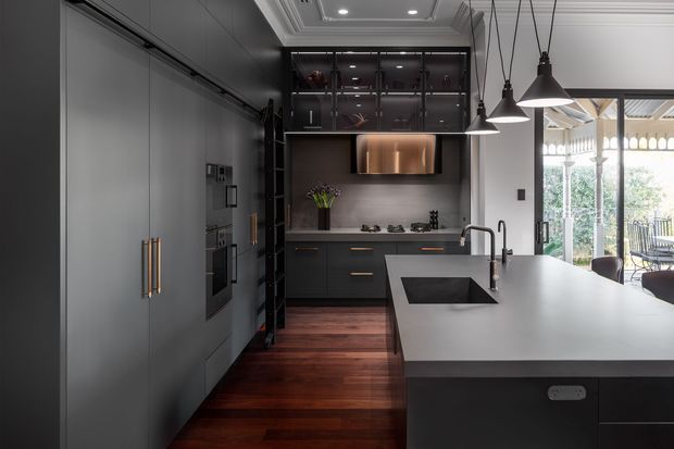 A sophisticated contemporary kitchen with dark matte cabinetry, integrated appliances, a large island with a dark stone or composite benchtop, and a timber floor. Black-framed glass-fronted overhead shelving, warm timber accents, and sculptural pendant lights create a refined contrast. A bright glazed outdoor area is visible beyond the kitchen.