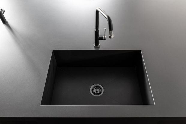 Close-up product-style view of a minimalist black undermount kitchen sink integrated into a dark grey benchtop, with a tall black and brushed-metal gooseneck mixer tap and a partially visible matching dispenser at the upper left. The monochromatic composition emphasizes clean geometric lines and reflective metal details.