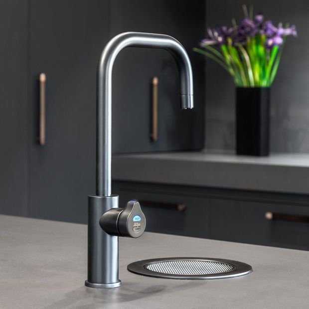 Close-up product-style view of a contemporary kitchen sink area featuring a tall brushed metal gooseneck tap with side lever, set into a grey stone benchtop. A round integrated sink drain or grate is visible in the foreground, with dark cabinetry, slim metallic handles, and a vase of flowers in the background.