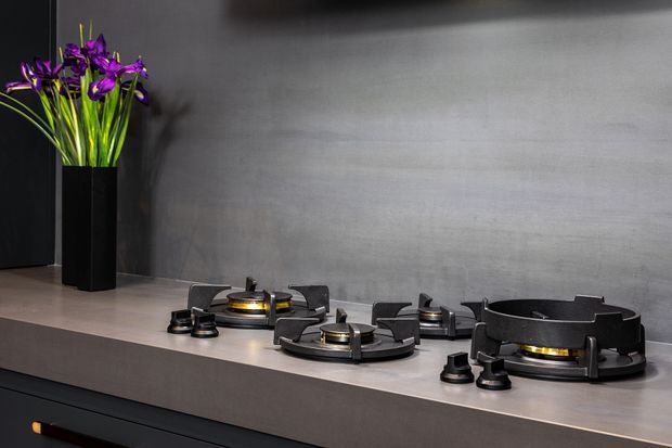 Close-up of a sleek modern black gas cooktop integrated into a dark grey stone or composite kitchen benchtop. The cooktop has multiple circular burners with metallic gold-toned burner components and substantial black pan supports. A tall black vase with vivid purple flowers sits at the back left, against a dark charcoal wall with subtle textured variation. The composition is moody, refined, and minimalist, focused on the cooking surface and surrounding built-in kitchen finish.