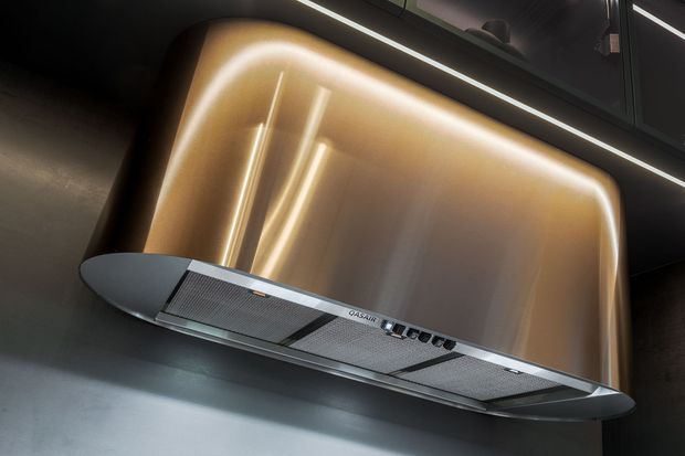 Close-up product-style image of a sleek stainless-steel rangehood mounted against a dramatic curved metallic splashback or wall panel. Integrated warm LED lighting outlines the rounded panel, with dark cabinetry and linear ceiling lighting above, creating a high-end contemporary kitchen setting.
