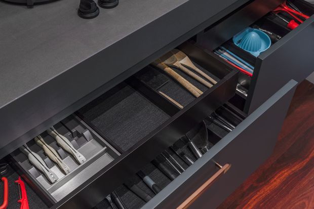 Close-up view of a dark, highly organised kitchen drawer system with custom internal dividers for cutlery, utensils, knives and other kitchen tools. The drawers have dark cabinetry, a grey countertop, metal organisers and a warm timber drawer handle, with reddish timber flooring visible at the side.
