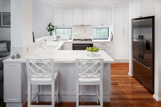 Bright, refined white kitchen with extensive built-in cabinetry, marble-look benchtops and splashback, a central island with seating for two, integrated cooking appliances, and a large stainless-steel refrigerator. Warm timber flooring and white lattice-back stools add contrast.
