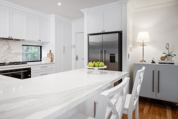 Bright, refined contemporary kitchen with extensive white paneled cabinetry, a large white stone island with waterfall edge, integrated stainless-steel appliances, marble-look splashback, and white cross-back bar stools. A grey sideboard and floor lamp sit in the adjoining area, with warm timber flooring adding contrast.
