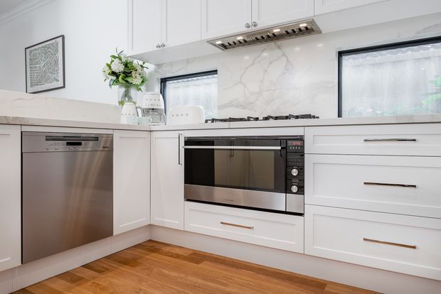 Bright, refined white kitchen with shaker-style cabinetry, integrated stainless-steel dishwasher, built-in oven and gas cooktop, marble-look splashback, and timber flooring. Two windows provide natural light, while slim metallic drawer pulls and a stainless-steel rangehood add contemporary detailing.