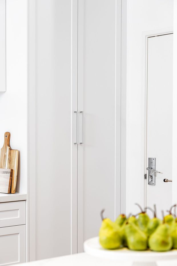 Bright, predominantly white interior showing tall shaker-style built-in cabinetry with slim vertical panel detailing and chrome handles. A white paneled door with a traditional metal lever latch is visible on the right, while a white drawer unit and timber chopping boards appear at the left. A blurred bowl of green pears sits in the foreground on a white counter or island.