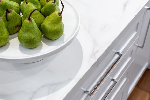 Close-up of a bright contemporary kitchen, showing a white marble-look benchtop with subtle grey veining, white shaker-style cabinetry, and long brushed metal drawer handles. A white serving bowl filled with green pears sits on the countertop; warm timber flooring is visible at the edge.