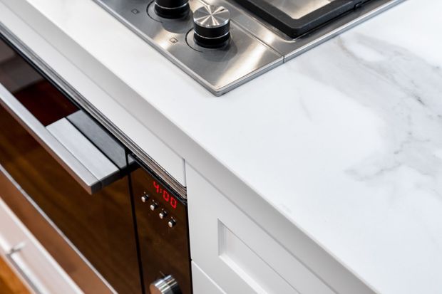 Close-up view of a contemporary kitchen benchtop featuring a white marble-look surface, stainless-steel gas cooktop, integrated oven, and white shaker-style cabinetry. Dark timber flooring is visible below.