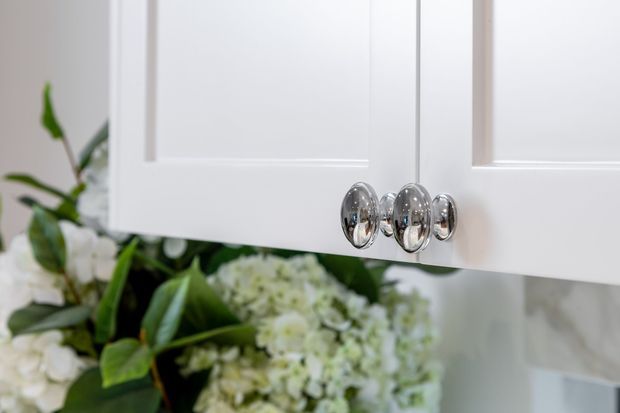 Close-up of white shaker-style built-in cabinetry with raised-panel doors and polished chrome knobs. Green leafy foliage and white flowers are visible behind the cabinetry, with a pale neutral wall in the background.
