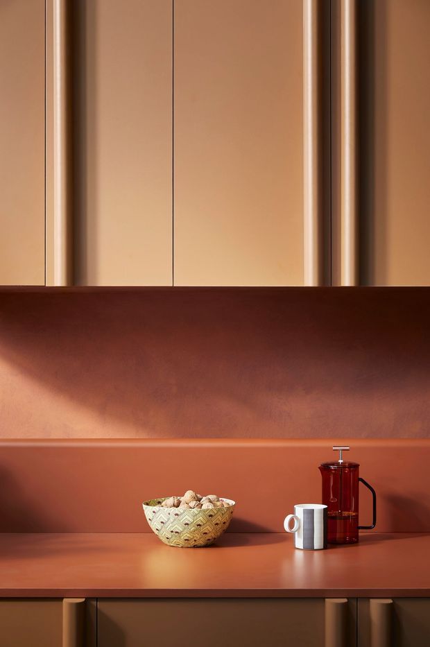 A close-up of a warm, monochromatic built-in kitchen or bar joinery composition. Terracotta and copper-brown cabinetry, a matching solid benchtop and backsplash, and vertically fluted or rounded cabinet framing create a refined sculptural backdrop. A woven bowl, white mug, and dark red glass coffee or tea carafe sit on the counter, adding small decorative accents. The image is tightly framed with no wider room context visible.
