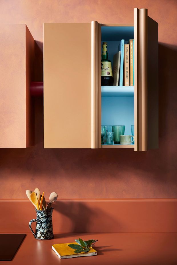 A close-up of a colourful wall-mounted storage cabinet or drinks cupboard above a kitchen benchtop. The cabinet has warm tan and terracotta doors with a bright blue interior, open shelving containing bottles, books and glassware, and rounded vertical timber-like edges. Below, a terracotta benchtop holds a patterned utensil jug and a book with a leafy branch. The image has a carefully composed, editorial product-photography style with strong warm-and-cool colour contrast.