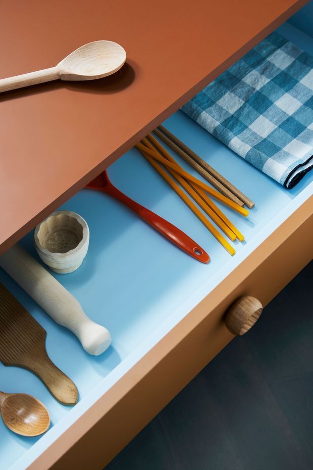 Close-up overhead view of an open built-in drawer or storage unit with a bright blue interior, containing wooden utensils, chopsticks or tools, a red-handled implement, wooden spoons, a small cup, and a folded blue-and-white checked cloth. The surrounding joinery is warm terracotta-brown, with a dark floor visible below. The image emphasizes organized storage and colourful material detailing rather than a full room.