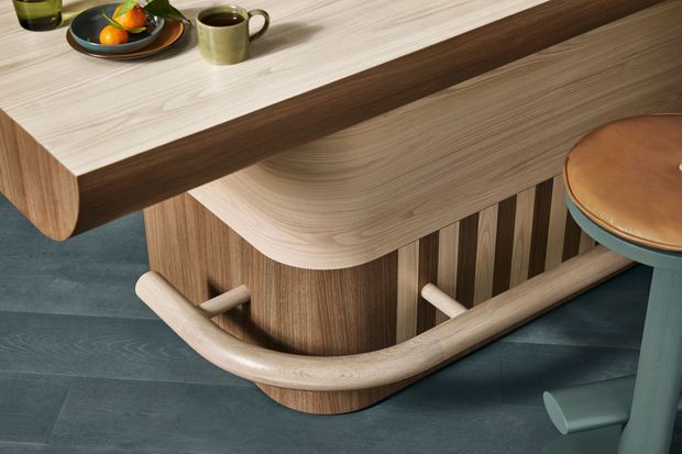 Close-up product-style view of a sculptural timber kitchen island or bar counter with rounded edges, layered light and dark wood finishes, curved integrated detailing, and vertical timber slats. A pale stone or wood-look benchtop holds a plate and mug, while a teal upholstered bar stool and dark flooring add contrast. The image emphasizes refined joinery and organic curved forms rather than a complete room.