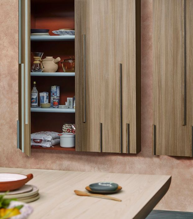 Close-up of a warm-toned kitchen or dining area featuring tall vertical-grain timber-look wall cabinetry with long slim handles. One cabinet is open, revealing crockery, pantry goods and serving vessels against a dark reddish interior. A pale timber dining table with ceramic dishes and wooden cutlery occupies the foreground, set against a textured pink-beige wall.