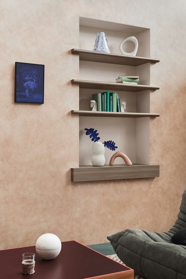 A styled living room vignette featuring a wall-mounted open shelving unit with integrated recessed backing and dark wood shelves, displaying books, vases, and sculptural objects. The surrounding wall has a warm textured blush plaster finish. A dark modular sofa and burgundy coffee table occupy the foreground, with a framed artwork mounted to the left. The composition is contemporary and curated, with warm earthy tones and sculptural decor.