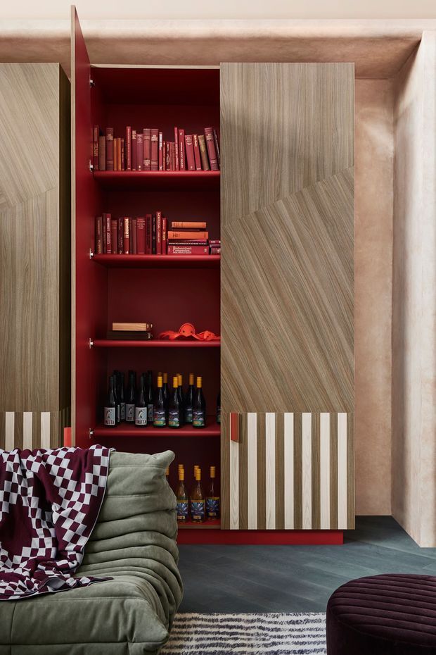 A richly styled living room featuring a tall built-in storage and display cabinet with deep burgundy-red interiors, open shelves of books and wine bottles, and patterned wood-grain doors with decorative striped lower panels. A plush olive-green sofa with a burgundy-and-white patterned throw occupies the foreground, alongside a dark burgundy ottoman and textured rug. The room has dark flooring and warm plaster-toned walls, creating a bold, eclectic atmosphere.