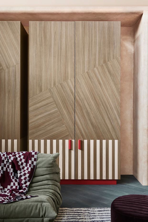A richly detailed contemporary interior featuring a large built-in timber wardrobe or storage wall with geometric grain-patterned doors and contrasting vertical slatted lower panels. The joinery sits against a warm plaster or stone-toned wall and includes small red handles and a red plinth. In the foreground are a deep olive-green upholstered sofa, a burgundy-and-white patterned throw, a dark burgundy ottoman, and a striped rug over dark flooring.
