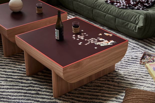 Close-up interior view of a contemporary living room centred on two low rectangular timber coffee tables with dark burgundy inset tops and thin red edging. The tables sit on a striped area rug beside a deep olive-green sofa. Board-game pieces, drinks, a bottle and decorative objects are arranged across the tables, creating a relaxed lounge setting.