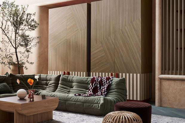 A richly styled living room featuring a deep olive-green modular sofa, sculptural timber coffee table, patterned throw, and woven ottoman. The room is defined by large decorative timber wall panels with geometric grain patterns, vertical timber screening, warm plaster-toned surfaces, and a tall indoor tree. The palette is earthy and muted with layered natural textures and strong built-in timber detailing.