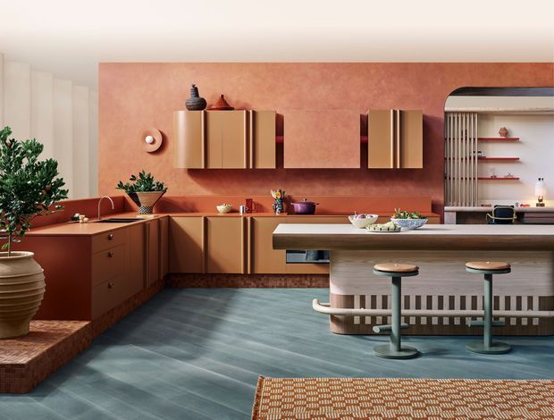 A richly styled contemporary kitchen with terracotta plaster walls, warm brown timber cabinetry, a long central island with integrated seating, and a curved open shelving and bar area. The room features a dark blue-grey floor, sculptural pendant lighting, patterned textiles, plants, and carefully curated decorative objects, creating a bold retro-inspired atmosphere.