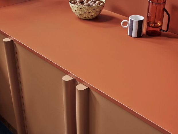 Close-up detail of warm terracotta-orange kitchen or furniture cabinetry with a matching solid countertop and distinctive rounded vertical door pulls. A ceramic bowl, striped mug, and glass coffee maker sit on the surface, emphasizing the rich monochromatic palette and smooth contemporary finish.
