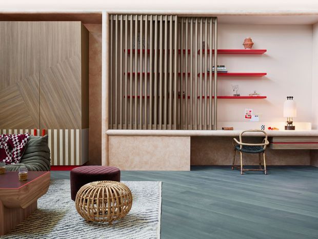 A stylish open-plan living and work area with a built-in desk, shelving, and a sculptural vertical timber screen. The room features warm textured plaster and timber joinery, vivid red shelving accents, a blue-grey timber floor, a patterned rug, and eclectic contemporary furnishings including a curved sofa, ottoman, woven coffee table, and cane chair.