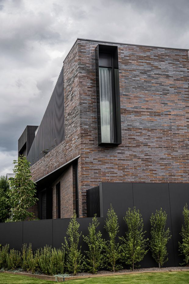 Exterior architectural photograph of a contemporary multi-level urban residence. The building has a strong cubic form, dark metal cladding and fencing, narrow floor-to-ceiling windows with black frames, and extensive variegated brown, grey, and muted terracotta brickwork. The foreground contains structured planting and a lawn under an overcast sky.