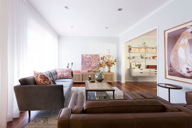 Bright, spacious living room with floor-to-ceiling sheer curtains, white walls and ceiling, timber flooring, and a mix of brown leather and grey upholstered sofas. A low timber coffee table, patterned rug, artwork, decorative objects, and a built-in shelving and cabinetry zone visible through the wide opening create a refined mid-century-inspired interior.