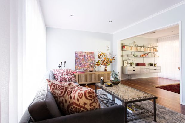 Bright, refined living room with a large dark upholstered sofa, patterned red and cream cushions, a long black coffee table, timber flooring, and a timber sideboard. A wide opening leads to an adjoining dining or sitting area with built-in shelving, books, and display objects. White walls, recessed ceiling lights, and abundant daylight create an airy contemporary interior.