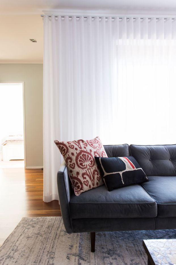 Bright living room featuring a large dark charcoal sectional sofa with patterned cushions, sheer white floor-to-ceiling curtains, pale neutral walls, timber flooring, and a muted patterned area rug. A doorway opens toward a bedroom in the background.