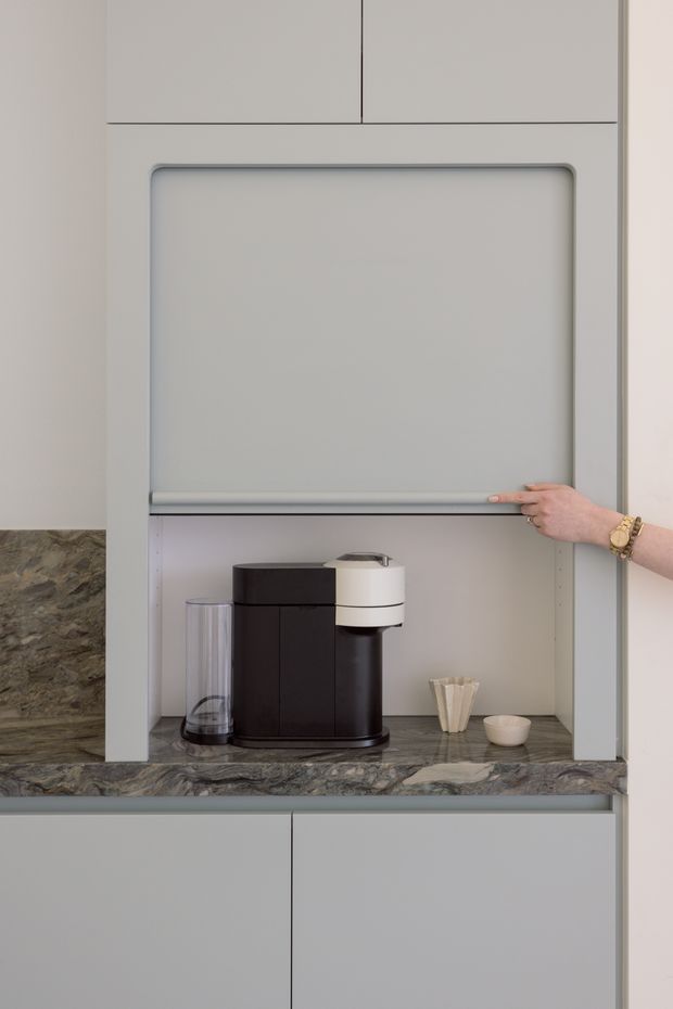 Close-up of a minimalist built-in coffee station within pale grey cabinetry. A raised roller door conceals the appliance nook, which has a dark veined stone benchtop, black and white coffee machine, clear water container, coffee filters and a small cup. The surrounding cabinetry is seamless and handleless, with a clean contemporary appearance.