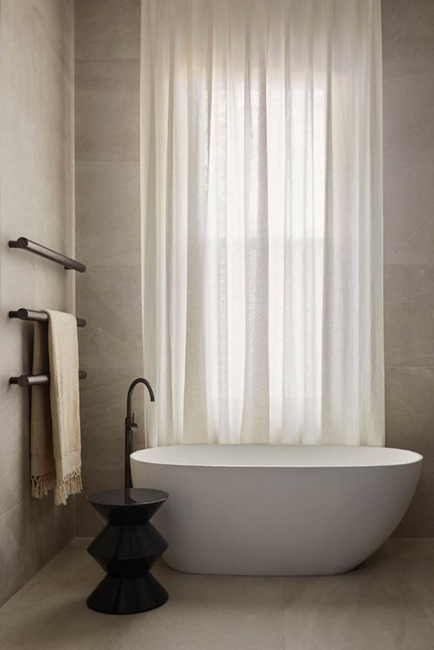 A serene, minimalist bathroom featuring a freestanding white oval bathtub positioned in front of tall sheer white curtains. Large-format warm grey-beige stone or porcelain tiles cover the walls and floor. Dark bronze or black towel rails and a sculptural black side table with a dark freestanding tap provide contrast.