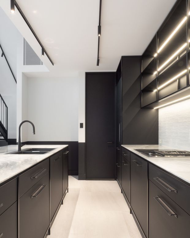 A sleek contemporary galley kitchen viewed down the central aisle. The room features extensive matte-black cabinetry on both sides, pale stone benchtops, a white brick-look tiled splashback, integrated linear LED lighting, black track lighting, a dark central door, and a black gooseneck tap over a double sink. A black-framed glazed stair enclosure is visible at the left, while the overall palette is monochromatic with strong clean-lined detailing.