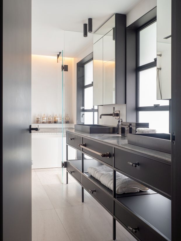 A bright, contemporary bathroom viewed from the vanity area, featuring a long dark charcoal double vanity with open shelving, drawers, two rectangular vessel basins, wall-mounted taps, and a pale stone benchtop. Large black-framed windows and mirrored wall cabinets reflect the room. A frameless glass shower screen separates the vanity from a freestanding white bathtub, with warm concealed lighting adding a soft glow.