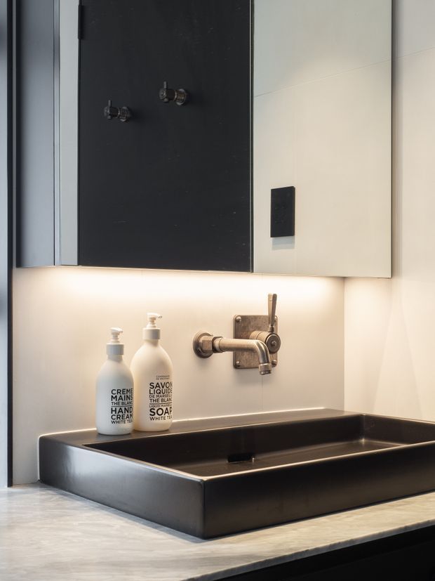 Close-up of a contemporary bathroom vanity featuring a large matte black rectangular vessel basin set on a pale stone benchtop, with wall-mounted brushed metal tapware, pale tiled walls, and mirrored cabinetry above. Integrated warm under-cabinet lighting illuminates the basin and countertop.