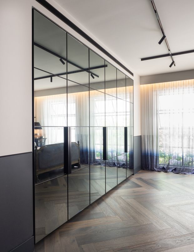 Bright contemporary interior with a full-height mirrored glass wall framed in slim black metal, reflecting a sofa and large windows. The room has pale walls with dark lower wall panelling, sheer floor-to-ceiling curtains, black track lighting, and a grey-brown timber herringbone floor.