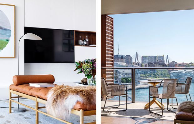 Split-view image showing a bright contemporary living room opening onto a balcony or terrace with expansive harbour and city views. The interior features white built-in cabinetry, a wall-mounted black television, a tan leather bench with a sheepskin throw, a sculptural floor lamp, and decorative shelving. The outdoor area has a small dining table and woven chairs beside full-height glazing, overlooking a marina and urban skyline.