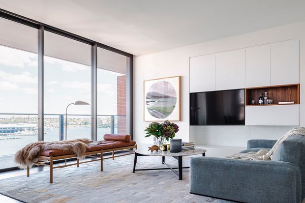 Bright contemporary living room with floor-to-ceiling glazing overlooking a waterfront marina. The room features white built-in media cabinetry with an open timber display niche, a large television, a blue-grey sofa, leather and fur-covered bench seating, a round coffee table, and a neutral patterned rug. Dark-framed glazing and a white ceiling create a clean, airy interior.