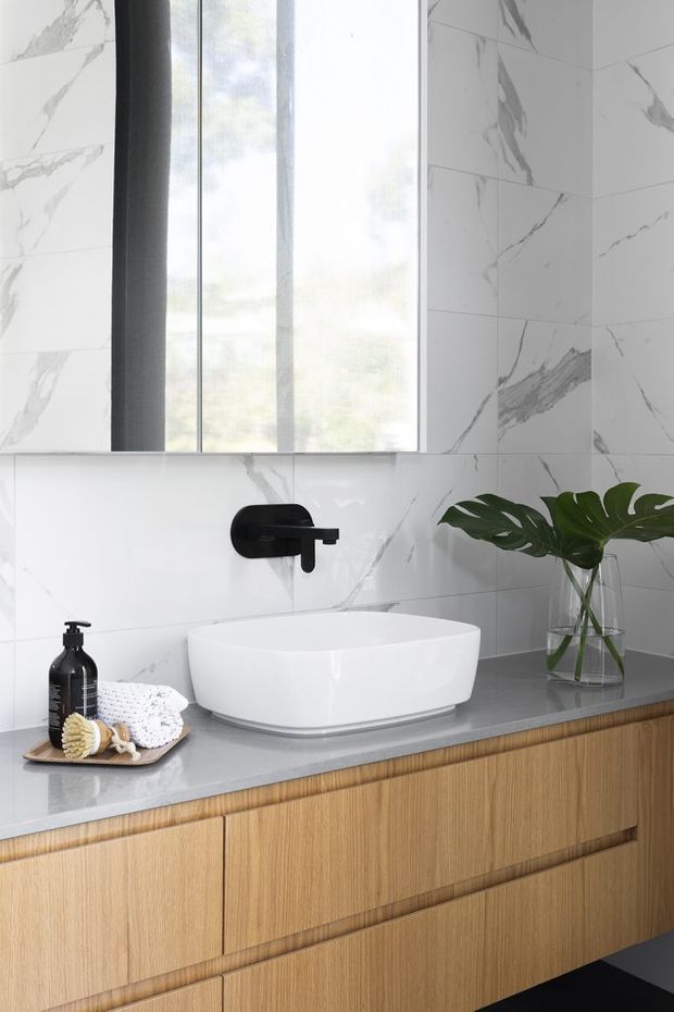 A contemporary bathroom vanity featuring a white vessel basin on a grey stone benchtop, warm timber drawer cabinetry, matte black wall-mounted tapware, and large white marble-look wall tiles with grey veining. A tall window provides natural light, with a small plant and toiletries on the counter.