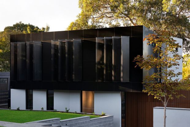 A contemporary multi-level residential facade with a dark, predominantly black upper volume and projecting vertical metal fins across the windows. The lower level combines white rendered wall panels, dark glazing, a warm timber-toned entry or feature panel, and vertical timber screening. Neatly landscaped lawns, paved steps, a low rendered boundary wall, and mature trees frame the building.